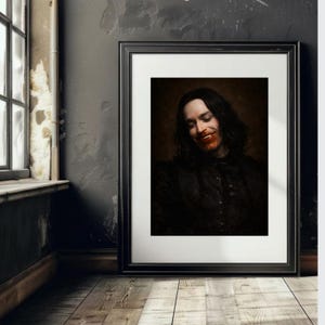 May include: Framed portrait of a person with long dark hair and a black outfit, smiling with red around the mouth. The portrait is in a black frame, leaning against a dark grey wall, with a wooden floor.