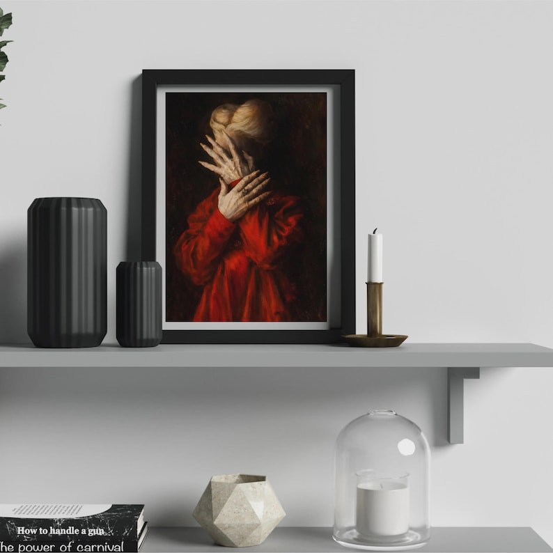 May include: Framed artwork featuring a person in a red garment, hands covering the face. The painting has a dark, moody aesthetic. The artwork is displayed on a gray shelf with black vases, a candle, and a decorative object.