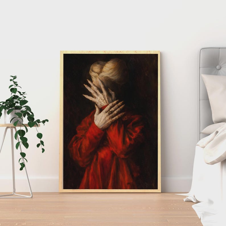 May include: Framed artwork featuring a person in a red garment, hands covering the face. The artwork has a dark background and a gold-colored frame. The person's hair is light-colored.