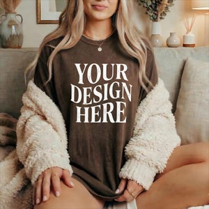 May include: A brown t-shirt with the text "YOUR DESIGN HERE" in white. The model is wearing a cream-colored, fuzzy jacket. The image is taken indoors, with a neutral background.