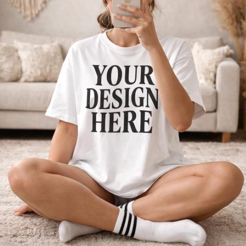 May include: A white t-shirt with the text "YOUR DESIGN HERE" in black block letters. The person is wearing white socks with black stripes. The person is sitting on a light-colored rug.