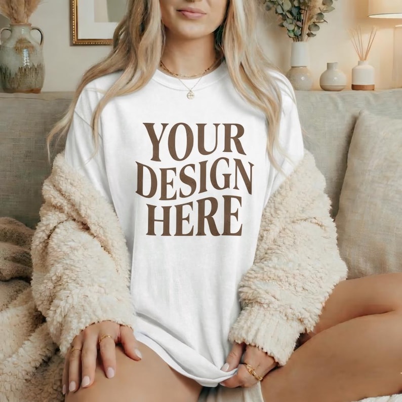May include: White t-shirt with the text "YOUR DESIGN HERE" in brown. The model is wearing a cream-colored, fuzzy cardigan. The model is also wearing gold jewelry.