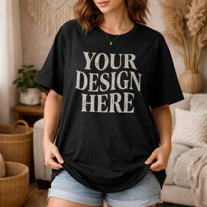 May include: Black t-shirt with the text "YOUR DESIGN HERE" in a distressed, off-white font. The shirt is short-sleeved and appears to be made of a soft material. The model is wearing blue denim shorts.