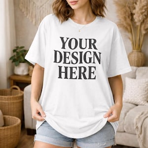 May include: White t-shirt with the text "YOUR DESIGN HERE" in a bold, black font. The shirt is being held up by a person. The person is wearing denim shorts. The background includes a woven basket and decorative plants.