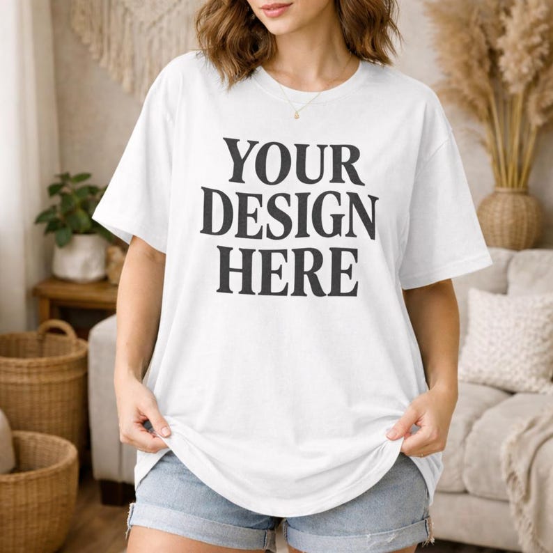 May include: White t-shirt with the text "YOUR DESIGN HERE" in a bold, black font. The shirt is being held up by a person. The person is wearing denim shorts. The background includes a woven basket and decorative plants.