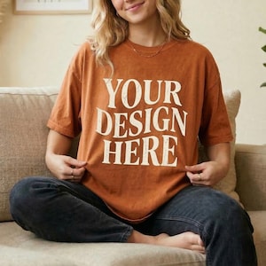 May include: A rust-colored t-shirt with the text "YOUR DESIGN HERE" in a bold, vintage-style font. The shirt is worn by a person sitting on a beige couch, wearing dark blue jeans. The overall aesthetic is casual and relaxed.