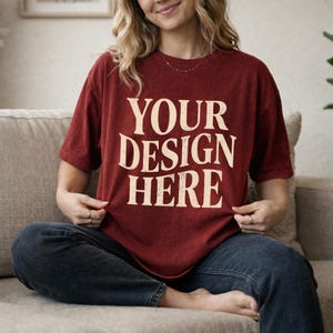 May include: A maroon t-shirt with the text "YOUR DESIGN HERE" in a distressed, off-white font. The shirt is worn with blue jeans. The model is sitting on a beige couch.