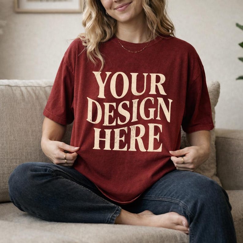 May include: A maroon t-shirt with the text "YOUR DESIGN HERE" in a distressed, off-white font. The shirt is worn with blue jeans. The model is sitting on a beige couch.