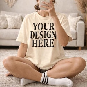 May include: Cream-colored t-shirt with the text "YOUR DESIGN HERE" in bold black letters. The shirt is a relaxed fit and appears to be made of soft material. The person is wearing white socks with black stripes.