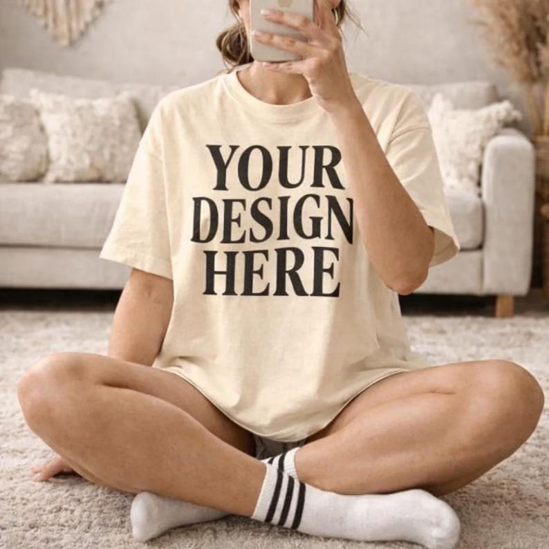 May include: Cream-colored t-shirt with the text "YOUR DESIGN HERE" in bold black letters. The shirt is a relaxed fit and appears to be made of soft material. The person is wearing white socks with black stripes.