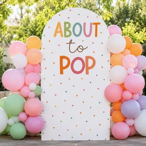 May include: A white arched backdrop featuring the text ABOUT to POP in colorful letters, decorated with a pattern of small multicolored dots. The backdrop is framed on both sides by a dense arrangement of balloons in shades of pink, orange, white, green, and lavender.