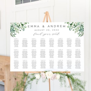 Op de afbeelding: Een zitplaatskaart voor een bruiloft met een groen en wit bloemenontwerp. De kaart staat op een houten ezel en zegt "Emma & Andrew August 20, 2022 Find your seat".