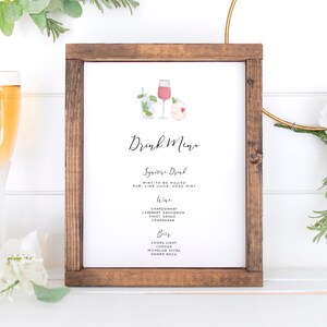 May include: A wooden framed sign with a white background and the text "Drink Menu" in black calligraphy. The sign features two watercolor illustrations of cocktails, one with a pink drink and the other with a green drink. The sign is sitting on a white surface with two champagne flutes and a floral arrangement.