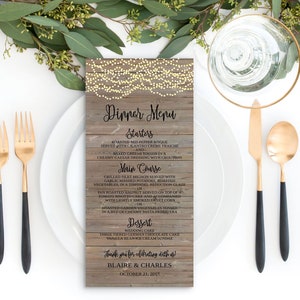 Puede incluir: Un men&uacute; de boda r&uacute;stico de madera con una cadena de luces, que enumera el men&uacute; de la cena para una recepci&oacute;n de boda. El men&uacute; incluye aperitivos, plato principal, postre y un mensaje de agradecimiento a los invitados. El texto dice: "Dinner Menu", "Starters", "Roasted Red Pepper Bisque", "Served with Cilantro Creme Fraiche", "and", "Mixed Greens Tossed in a", "Creamy Caesar Dressing with Croutons", "Main Course", "Grilled Filet Mignon Served with", "Garlic Mashed Potatoes, Roasted", "Vegetables, in a Zinfandel Reduction Glaze", "or", "Pan Roasted Halibut Served on Top of a", "Tomato Risotto Cake and Accompanied", "with Lightly Smoked Sweet Corn", "Roasted Garden Vegetables Tossed", "in a Bed of Creamy Pasta Primavera", "Dessert", "Wedding Cake", "Three Tiered German Chocolate Cake", "Vanilla Bean Ice Cream Sundae", "Thank you for celebrating with us!", "Blaire & Charles", "October 21, 2017".
