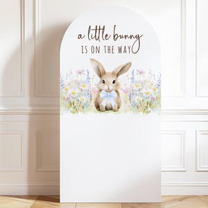 May include: White arched sign with the text "a little bunny is on the way." A watercolor illustration of a bunny with a blue bow tie is surrounded by wildflowers. The sign is a decorative element for a baby shower or nursery.