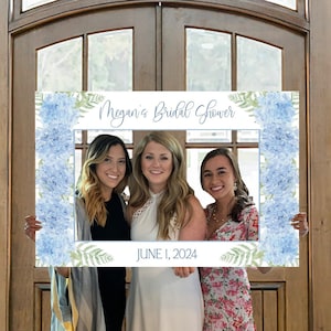 May include: A blue and white photo booth frame with a floral design and the text "Megan's Bridal Shower" and "JUNE 1, 2024". Three women are holding the frame and smiling at the camera.