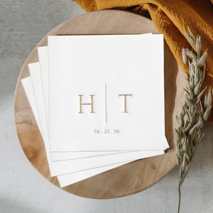 May include: A stack of white cards with gold lettering on a wooden surface. The cards have the initials "H" and "T" separated by a vertical line and the date "06.21.26".