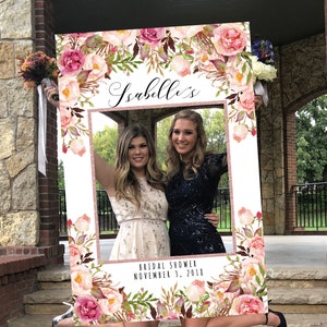 May include: A floral photo booth prop with a white frame and a pink and green floral design. The text "Isabelle's Bridal Shower November 3, 2018" is printed on the bottom of the frame.