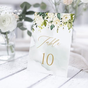 Pode incluir: Cartão de número de mesa floral branco com caligrafia dourada que diz "Mesa 10" em um fundo de aquarela verde claro.