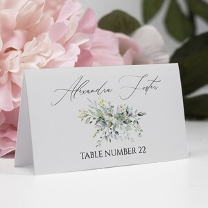 May include: White place card with a green floral design and the text "Alexandra Foster" and "Table Number 22".