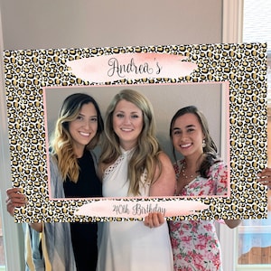 May include: A photo booth prop featuring a leopard print background with gold accents. The prop has a pink and white brushstroke design with the text "Andrea's 40th Birthday" written in black.