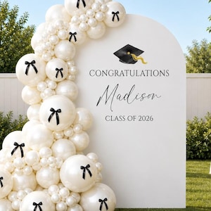 May include: A graduation party scene with a white arch decorated with white balloons and black bows. The arch features the text "Congratulations Madison Class of 2026" and a graduation cap graphic. White tables with floral arrangements are in the background.