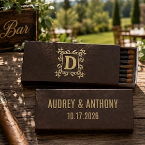May include: Two rectangular dark brown matchboxes with gold foil lettering. One box features a decorative frame with the letter D, while the other displays the text AUDREY & ANTHONY and the date 10.17.2026. The boxes contain matches with dark tips.