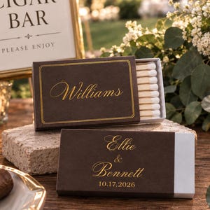 May include: Two rectangular matchboxes with dark brown covers and gold foil lettering. One box displays the name Williams, while the other features the names Ellie & Bennett with the date 10.17.2026. The boxes contain white-tipped wooden matches.