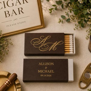 May include: Two rectangular dark brown matchboxes with gold foil lettering. One box features the initials AM in script, while the other displays the names ALLISON & MICHAEL and the date 09.19.2026. The open box reveals wooden matches with white tips.
