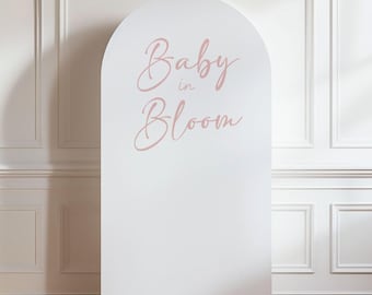 May include: A white, arched backdrop with the words "Baby in Bloom" in a soft pink script. The backdrop is set against a white wall with decorative molding and a wooden floor.