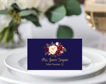 May include: A rectangular navy blue place card featuring a watercolour floral arrangement of white and dark red flowers. The text Mrs. Joanne Simpson is printed in gold script, followed by the text Table Number 22 in gold lettering.