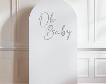 May include: A white, arched backdrop with the words "Oh Baby" in a soft green script. The backdrop stands on a wooden floor against a white wall with molding. The design is simple and elegant, suitable for a baby shower or gender reveal.
