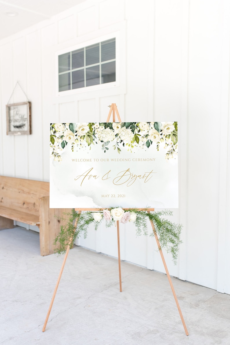May include: A white wedding welcome sign with green floral accents and gold lettering. The sign reads "Welcome to our wedding ceremony Ava & Bryant May 22, 2021". The sign is displayed on a wooden easel.