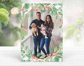 May include: A Christmas card featuring a family of three with a dog. The family is standing on a porch with a white railing. The image is framed with a watercolor wreath of green leaves and red berries.