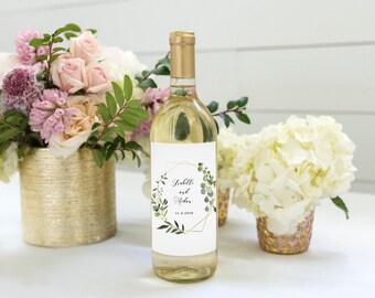May include: A clear glass wine bottle with a gold foil cap and a white rectangular label. The label features a gold geometric frame, green eucalyptus leaves, and the text "Isabelle and Asher 11.3.2018" in a black script and serif font.