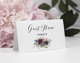 May include: A white folded place card featuring the black printed text Guest Name and TABLE 8, decorated with a watercolour illustration of dark red, blue, and green flowers and foliage at the bottom centre of the card.