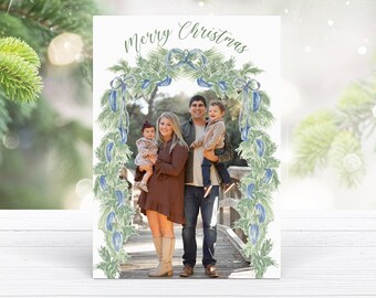 May include: A rectangular Christmas card featuring a watercolor illustration of a green pine garland arch with blue ribbons. The text Merry Christmas is printed at the top. A photograph of a family of three is centered within the arch.