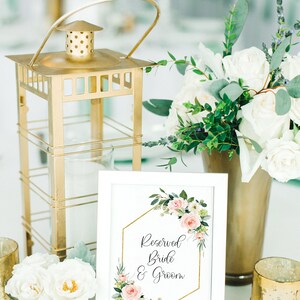 Puede incluir: Un cartel enmarcado en blanco con un marco hexagonal dorado y un dise&ntilde;o floral rosa dice "Reservado Novia & Novio". El cartel est&aacute; sobre una mesa con una linterna dorada, flores blancas y candelabros dorados.