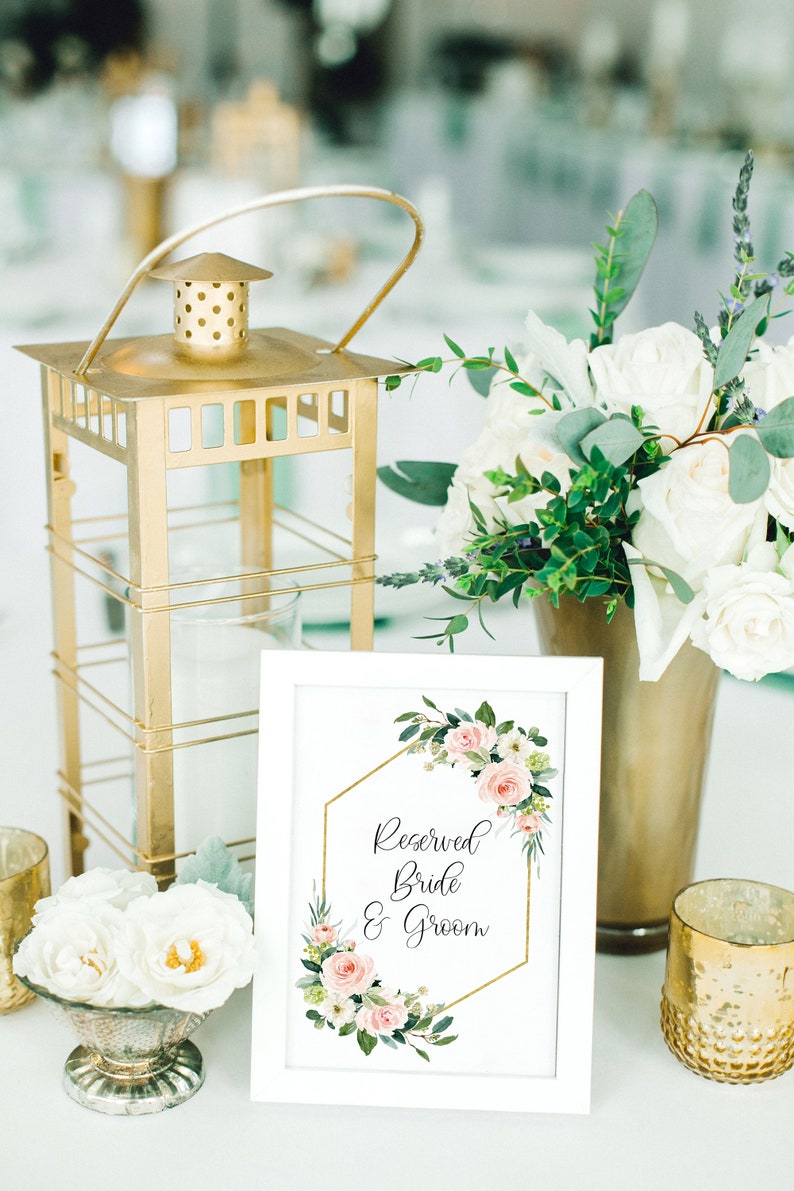 Puede incluir: Un cartel enmarcado en blanco con un marco hexagonal dorado y un dise&ntilde;o floral rosa dice "Reservado Novia & Novio". El cartel est&aacute; sobre una mesa con una linterna dorada, flores blancas y candelabros dorados.