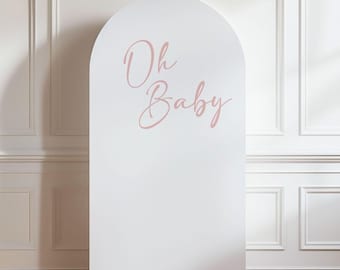 May include: A white, arched backdrop with the words "Oh Baby" in a soft pink script. The backdrop stands against a wall with white paneling and a wooden floor, creating a clean and elegant aesthetic, perfect for baby showers or gender reveal parties.