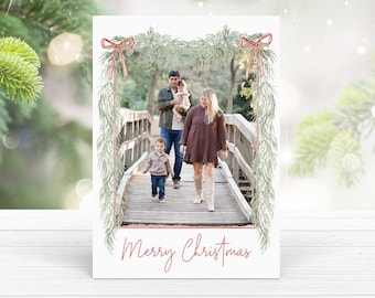 May include: A Christmas card showing a family of four walking on a wooden bridge. The parents are holding a baby and a toddler. The card has a green garland with red bows and the text "Merry Christmas" in red.