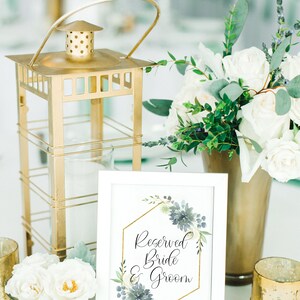 May include: A white sign with a gold geometric frame and blue watercolor flowers reads "Reserved Bride & Groom". The sign is on a table with a gold lantern, white flowers, and gold candle holders.
