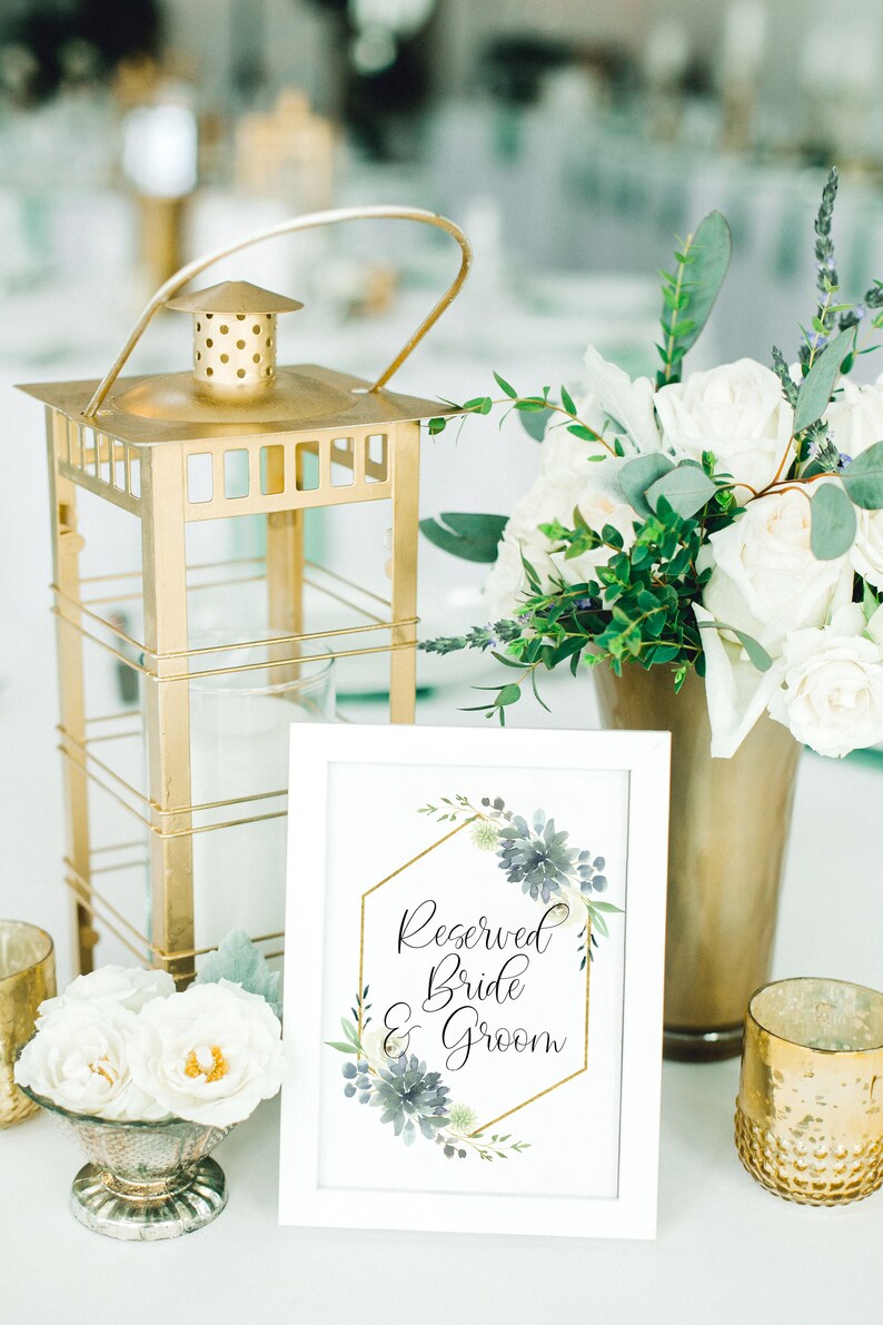 May include: A white sign with a gold geometric frame and blue watercolor flowers reads "Reserved Bride & Groom". The sign is on a table with a gold lantern, white flowers, and gold candle holders.