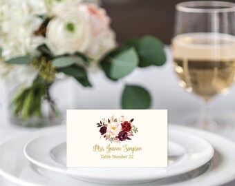 May include: A rectangular white place card featuring a floral illustration of cream, pink, and deep burgundy roses with green leaves. The text printed in gold script reads Mrs. Joanne Simpson and below it in gold text reads Table Number 22.