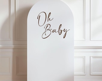 May include: A white, arched backdrop with the words "Oh Baby" in elegant brown script. The backdrop is set against a white wall with decorative molding, creating a clean and sophisticated look, perfect for baby showers or gender reveal parties.