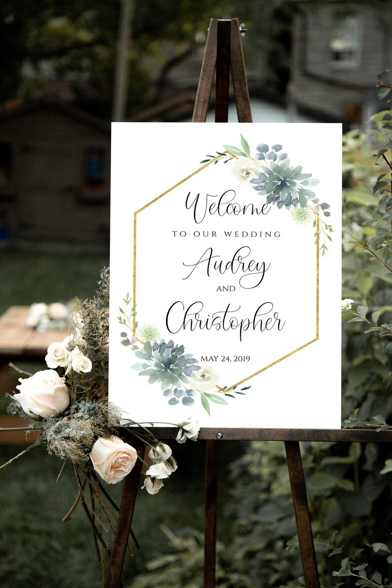 May include: Welcome to our wedding sign with a gold hexagonal frame and watercolor greenery. The sign reads "Welcome to our wedding Audrey and Christopher May 24, 2019".
