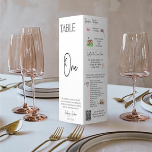 Op de afbeelding: Een wit rechthoekig bruiloftstafelbordje met het opschrift Table One in zwarte tekst, voorzien van een lijst met receptieactiviteiten, een I Spy-spel, een QR-code en de namen Audrey en Jackson met de datum 17-08-2024 op de voorzijde.