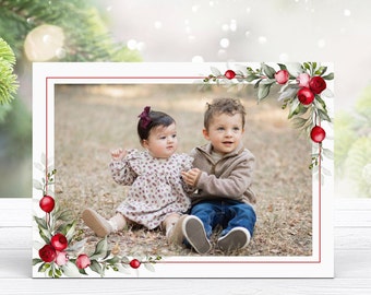 May include: A Christmas card featuring a watercolor illustration of red berries and green leaves surrounding a photo of two children. The children are sitting on the ground, smiling and looking up. The girl is wearing a floral dress and a red bow in her hair. The boy is wearing a brown sweater and blue jeans.