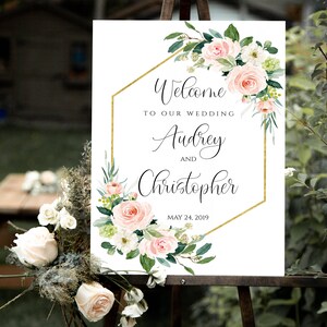 May include: A white wedding welcome sign with a gold geometric frame and pink and white floral accents. The sign reads "Welcome to our wedding Audrey and Christopher May 24, 2019".