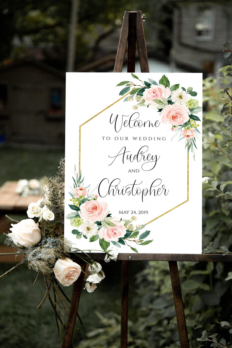 May include: A white wedding welcome sign with a gold geometric frame and pink and white floral accents. The sign reads "Welcome to our wedding Audrey and Christopher May 24, 2019".
